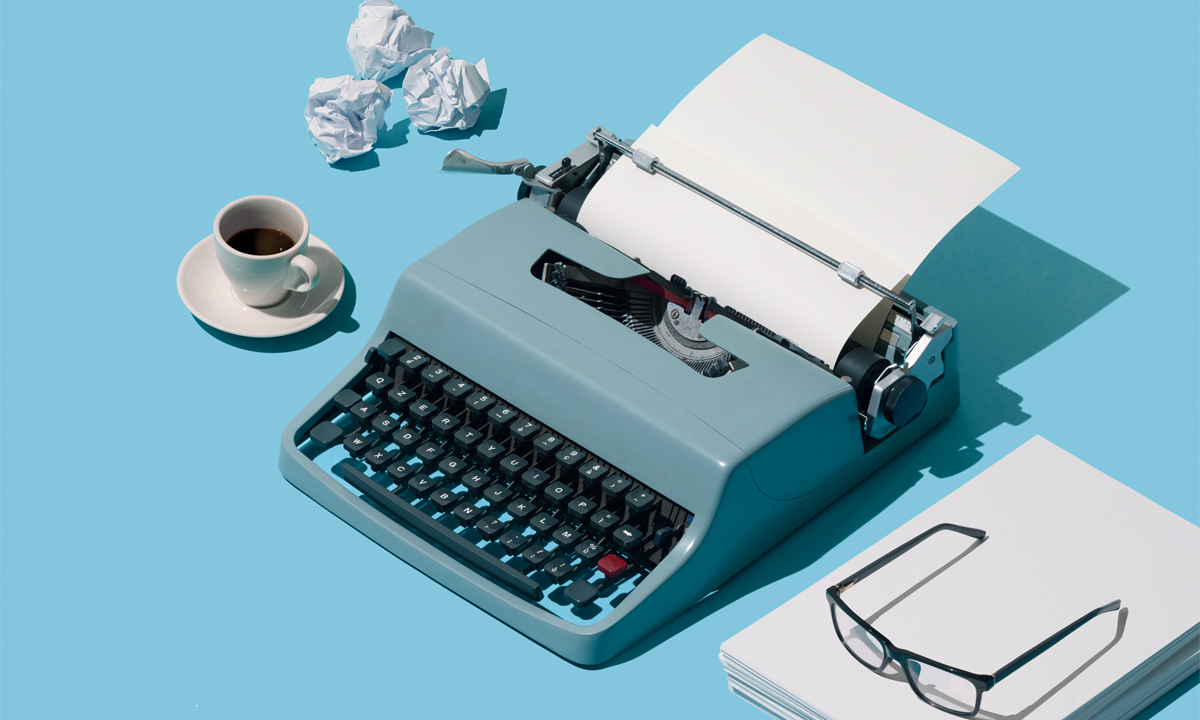 A table with a a typewriter, coffee cup, glasses and tossed paper. 
