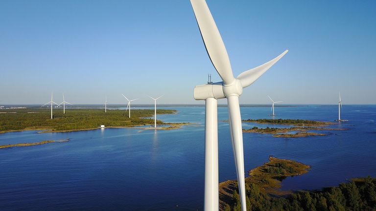 Several wind turbines are positioned over water, with more turbines in the distance against a backdrop of trees and a clear blue sky.